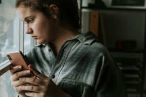 a teen girl holds her smartphone while looking out the window and thinking about the impact of social media on youth