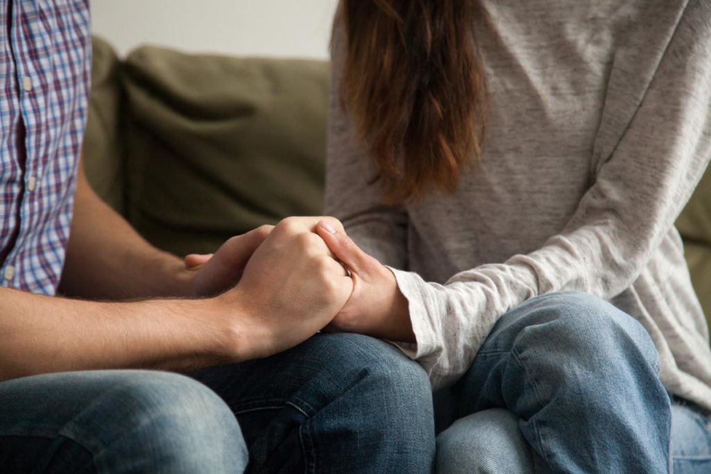 two people sit on a couch holding each others hands after understanding the connection between trauma and relationships