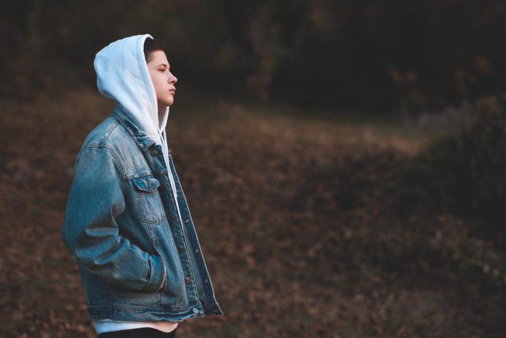 a man wearing a hoodie and jean jacket stares off in the distance while outside while also thinking about the signs of disruptive mood regulation disorder