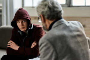 a teen boy wears a hoodie with his hood up while talking to a therapist about his risky behaviors