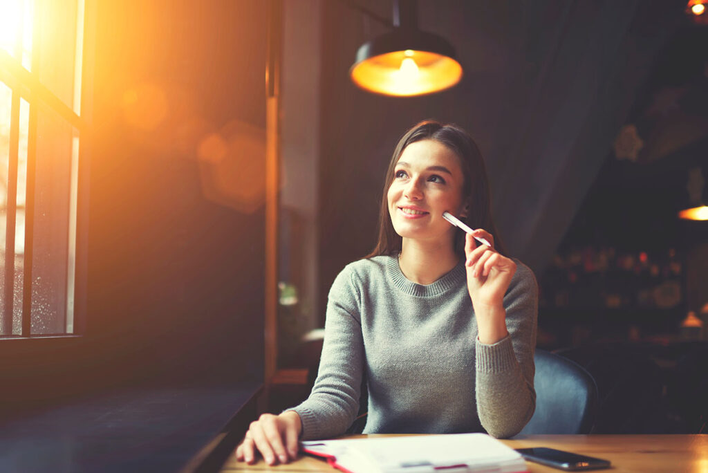 a woman sits at a desk with a pen to her face while she looks afar and thinks about how to practice mindfulness