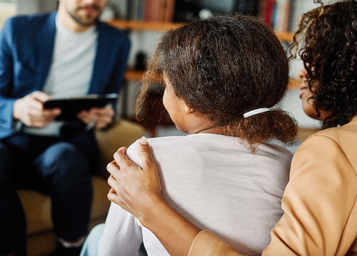 a mother uses her arm to hug her daughter who is sitting next to her while they talk to a therapist and learn how to healing from childhood trauma