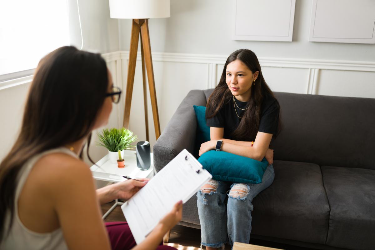 a therapist holds a clipboard and talks with her teen patient who is sitting on a couch about symptoms of bipolar disorder in teens