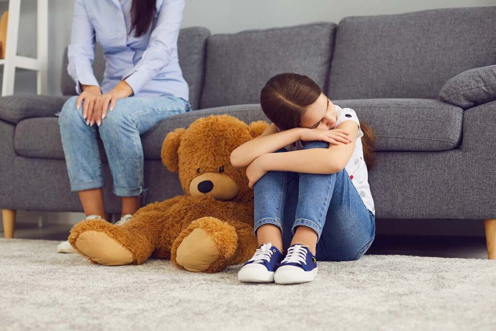 depressed young girl seated on floor of therapist's office next to large stuffed animal while her mother ask therapist, "How does childhood depression affect adulthood?"
