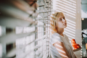 young woman leans against closed window shades staring into space as an example of depression rates in Los Angeles