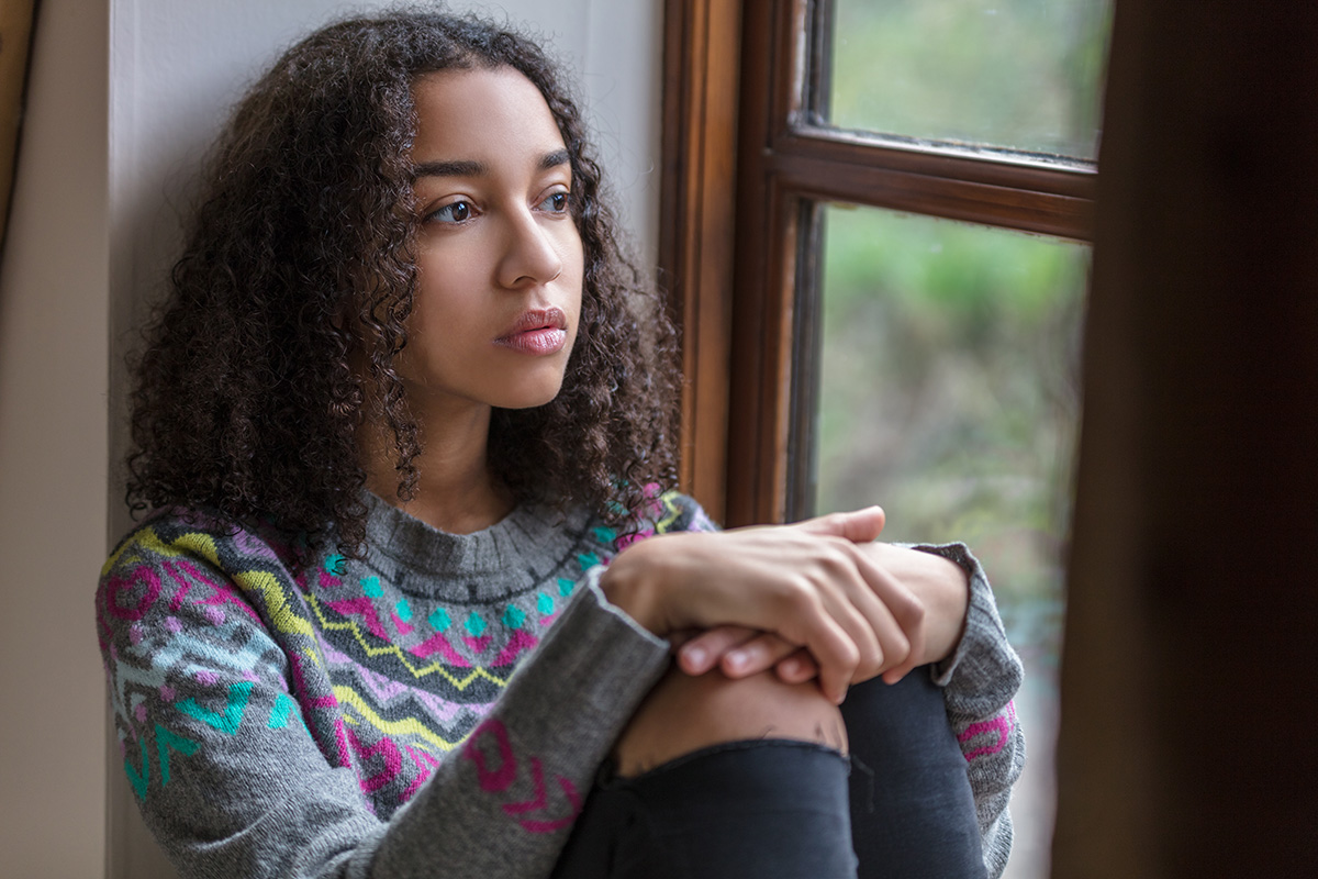 young girl sits by window looking sad and lonely as a result of the link between chldhood abuse and depression